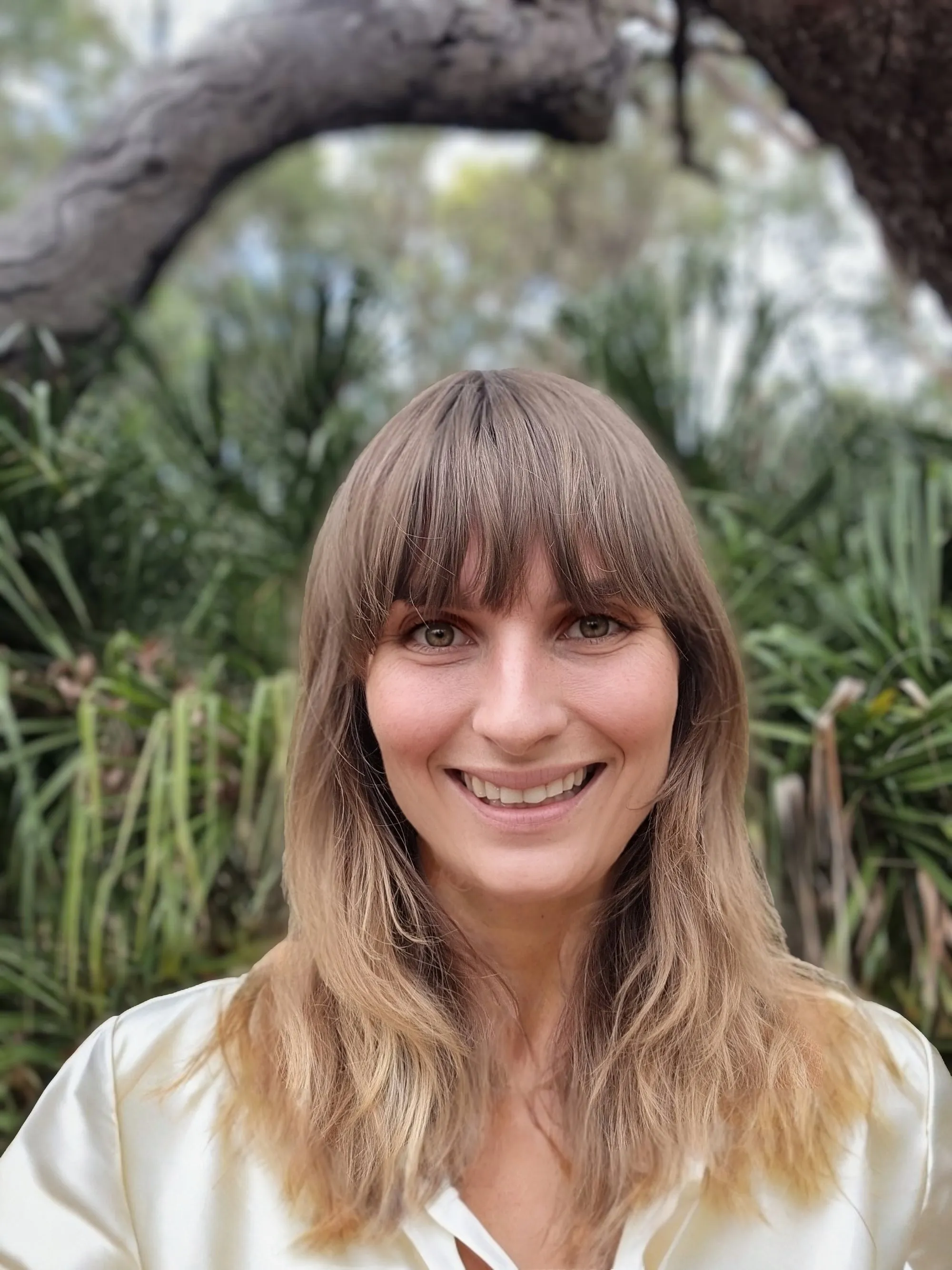Nikita Hall, smiling to camera, outdoors in front of coastal scrub and a low tree branch.